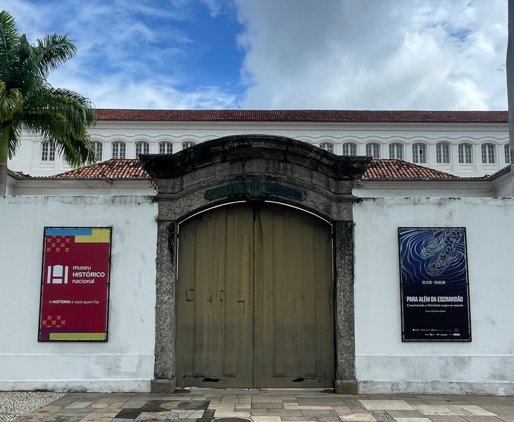 Entrance of the National Historical Museum featuring large wooden doors and two informative banners on either side. A palm tree is visible in the foreground against a partly cloudy sky.